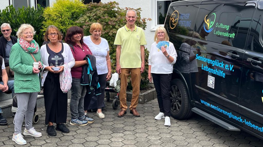 wurde gemeinsam mit einem herzensguten Spender ins Leben gerufen. Vor dem Gemeindehaus am Hellweg haben sich die Seniorinnen und Senioren zur Seniorentafel in Körne eingefunden. Eine Seniorin, ganz rechts im Bild, hält ein Bündel Lebensmittel-Gutscheine von ALDI in der Hand.