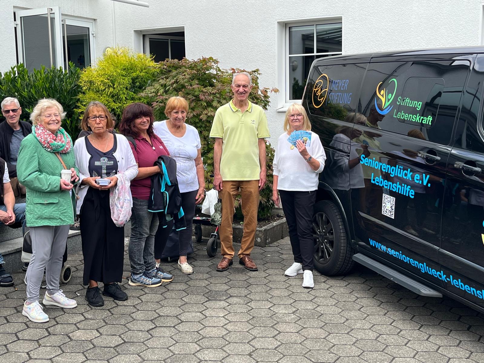 Am Lieferbus haben sich die bedürftigen Seniorinnen und Senioren zur Seniorentafel in der Gemeinde am Hellweg in Dortmund-Körne eingefunden. Eine Seniorin, ganz rechts im Bild, direkt neben dem Bus, hält ein Bündel Lebensmittel-Gutscheine in der Hand.
