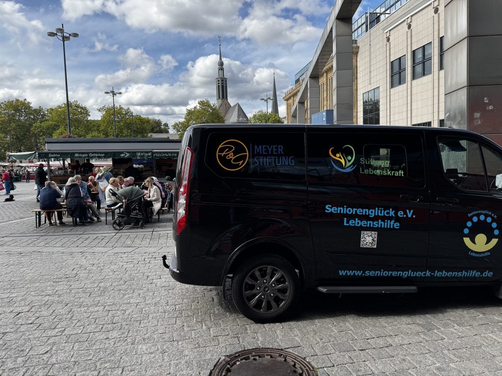 Der Seniorenglück e.V.-Bus vor dem Marktstand von Tevil's Fischwerk am Dortmunder Wochenmarkt mit den Aufschriften Seniorenglück e.V. Lebenshilfe - www.seniorenglueck-lebenshilfe.de Stiftung Lebenskraft P&G MEYER STIFTUNG