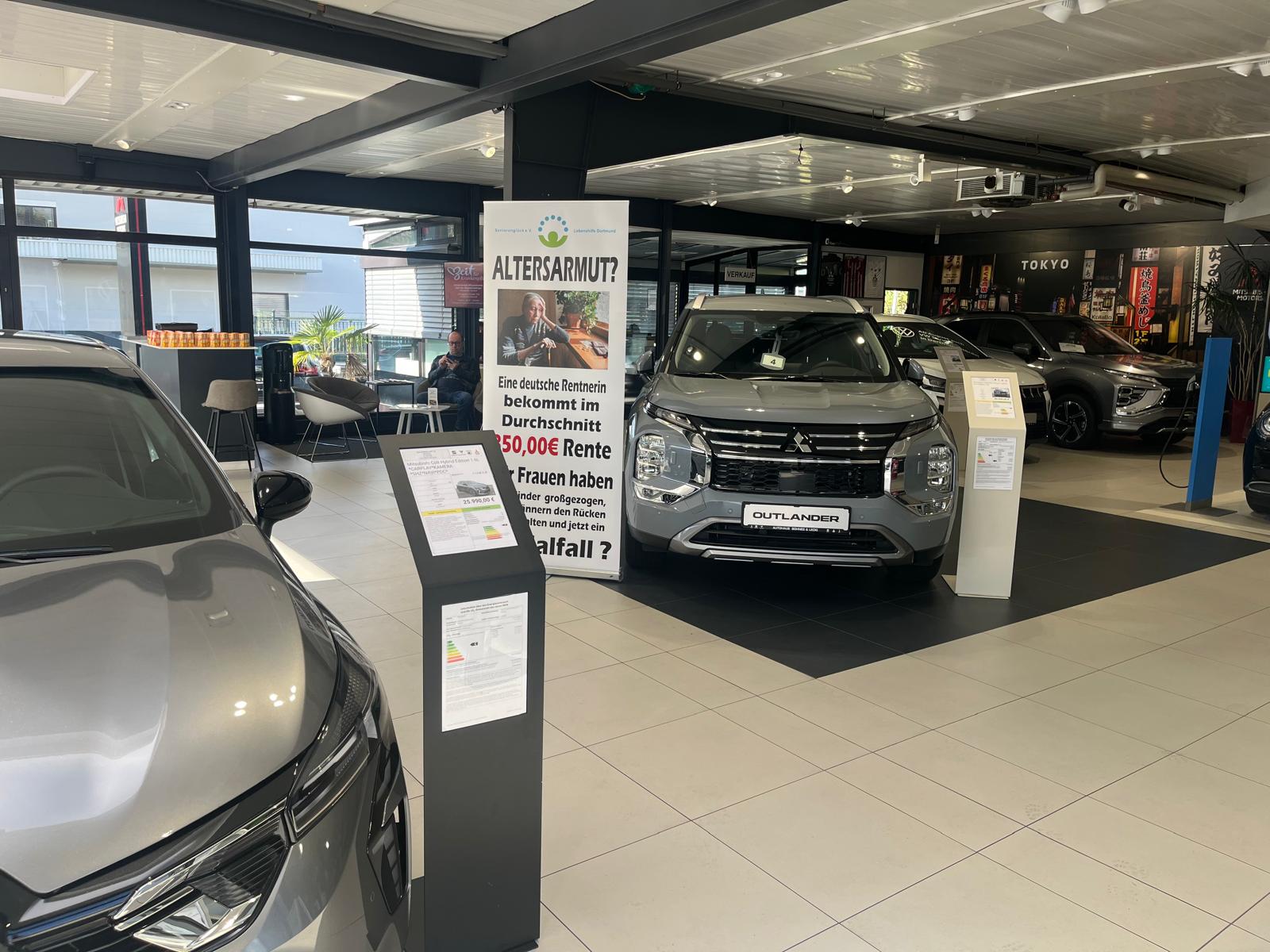 Ein Mitsubishi Outlander in Alpina Grau. Daneben steht ein großes Schild: Altersarmut? - Eine deutsche Rentnerin bekommt im Durchschnitt 850,00 € Rente Wir Frauen haben Kinder großgezogen, den Männern den Rücken frei gehalten - und jetzt ein Sozialfall?