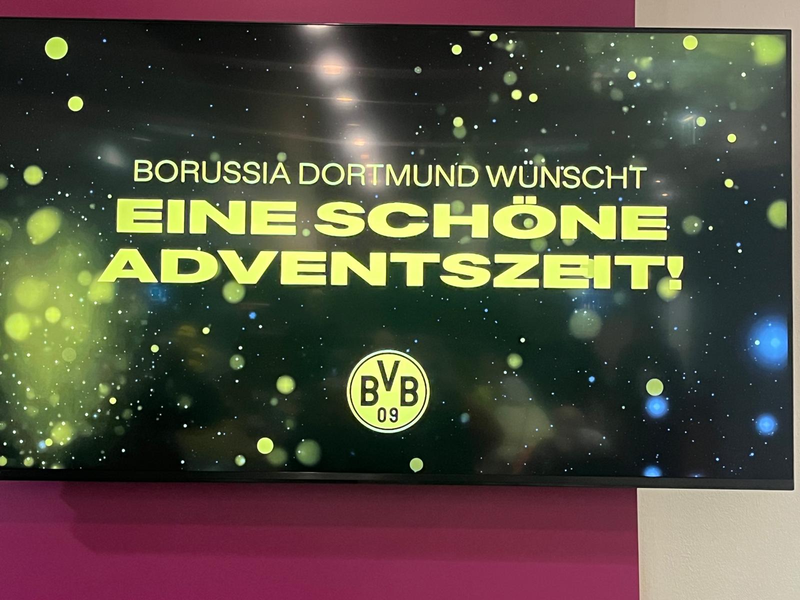 "Borussia Dortmund wünscht eine schöne Adventszeit!" steht auf einem großen Wandbildschirm in gelber Schrift. Darunter das BVB 09 Logo.