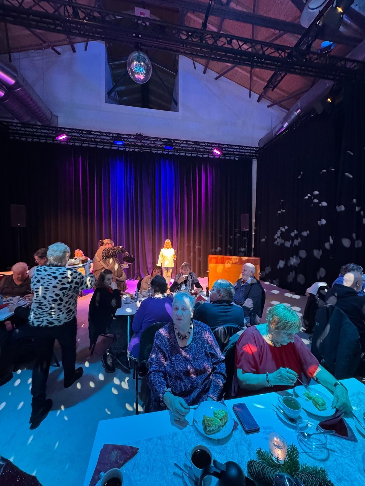 Frau Cornelia Sbosny steht auf der Bühne, bereit zur Fest-Ansprache, die Gäste genießen Kaffee und Kuchen.