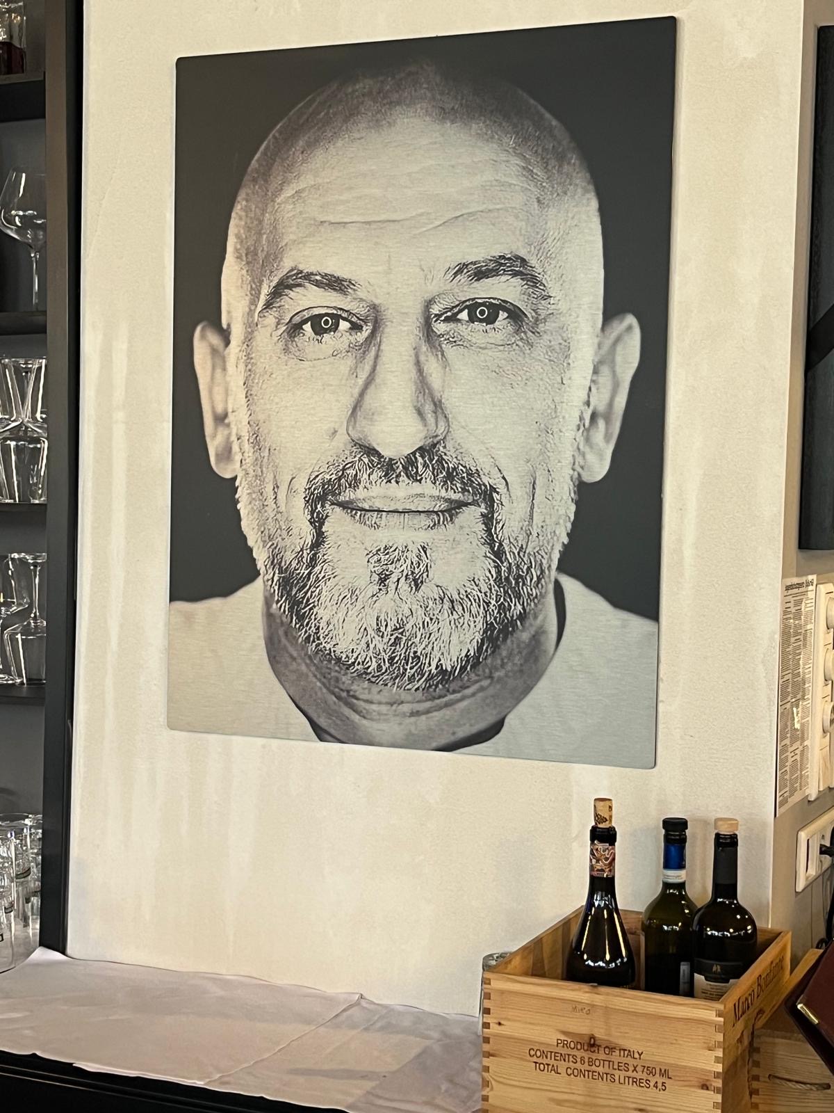 Ein Porträt von Bejan Sokoli, dem nach langer Krankheit verstorbenen früheren Besitzer des Ristorante Scuderia. Davor eine Holzkiste mit 3 Flaschen Wein und der Aufschrift "Product of Italy, contents 6 bottles x 750 ml, total contents litres 4,5"